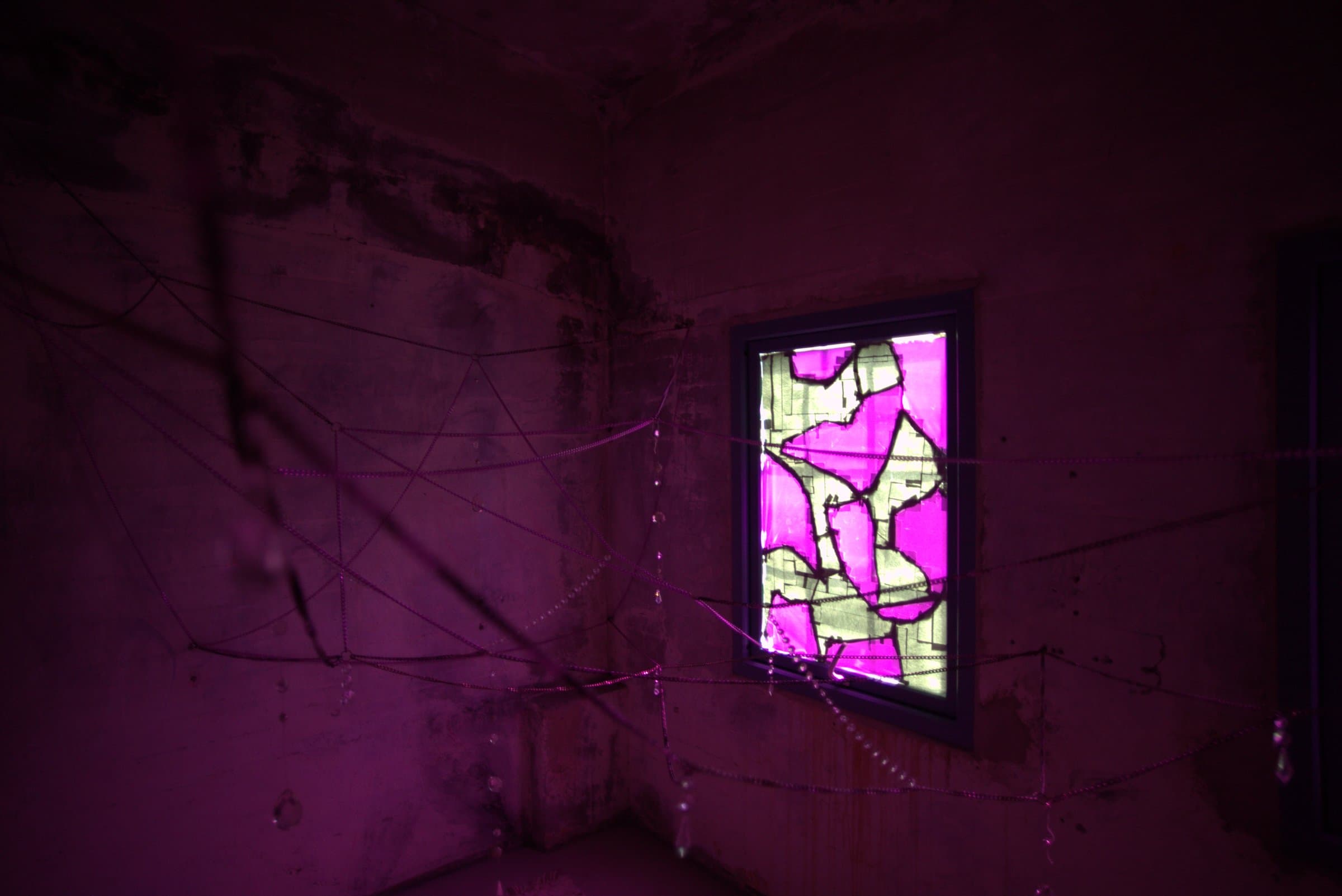 A magenta-lit stained-glass window in a concrete room corner, seen through a web of hanging beaded chains