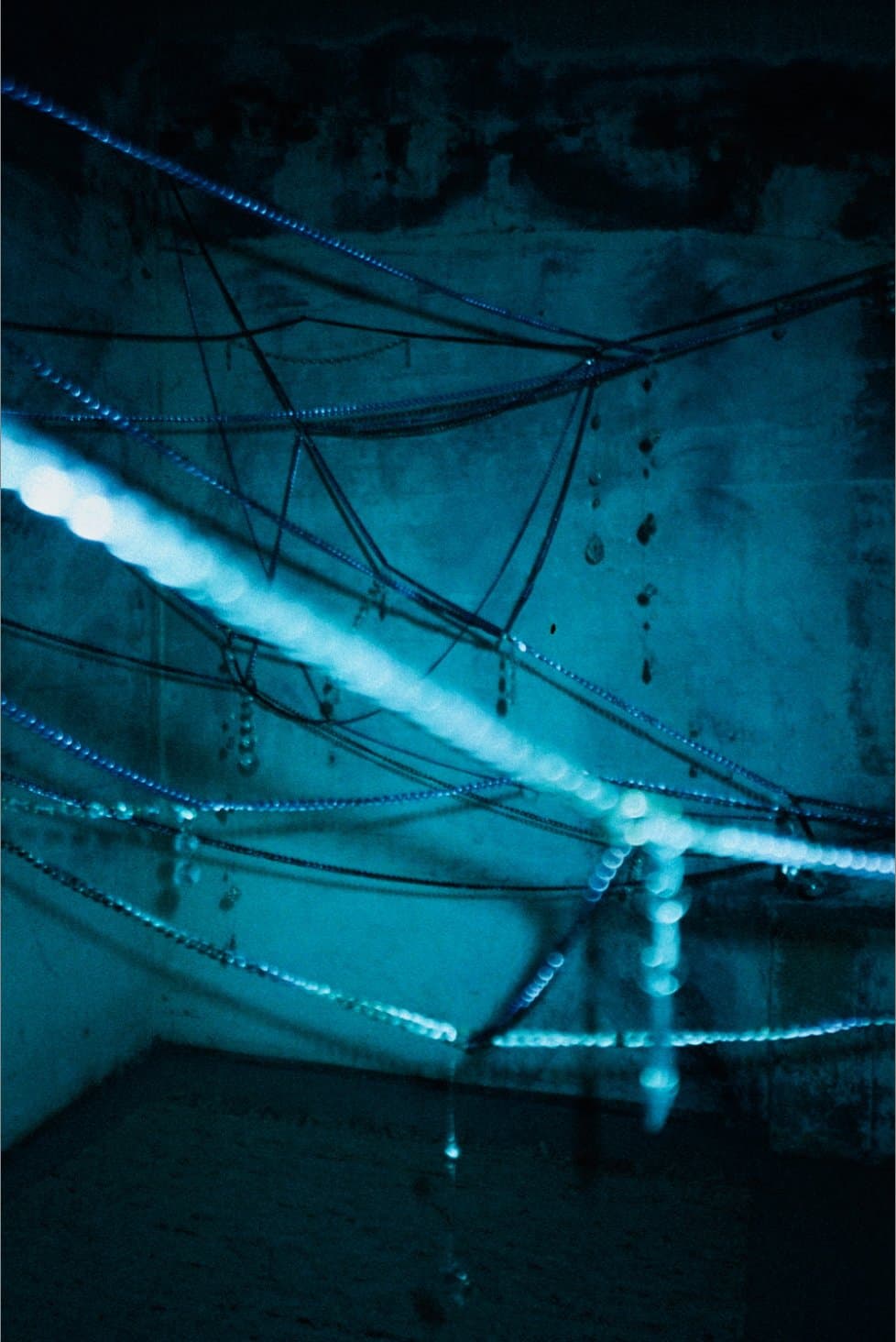 Blue-lit beaded chains strung across a concrete corner, drops catching the light against the bare wall