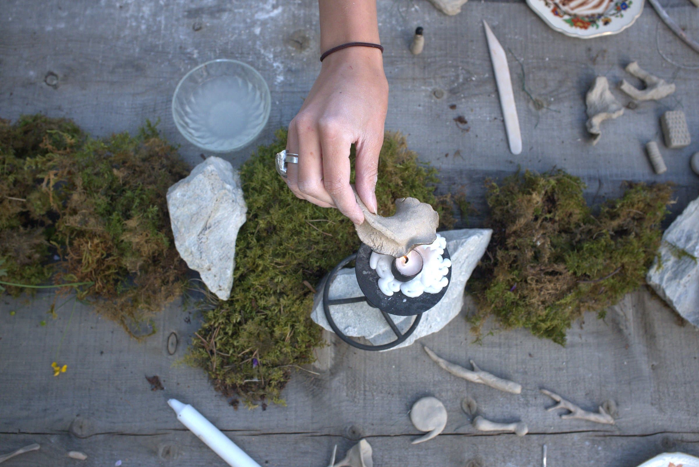 A hand placing a clay piece onto a candle holder during a sculpture workshop, surrounded by moss, stones, and small ceramic objects