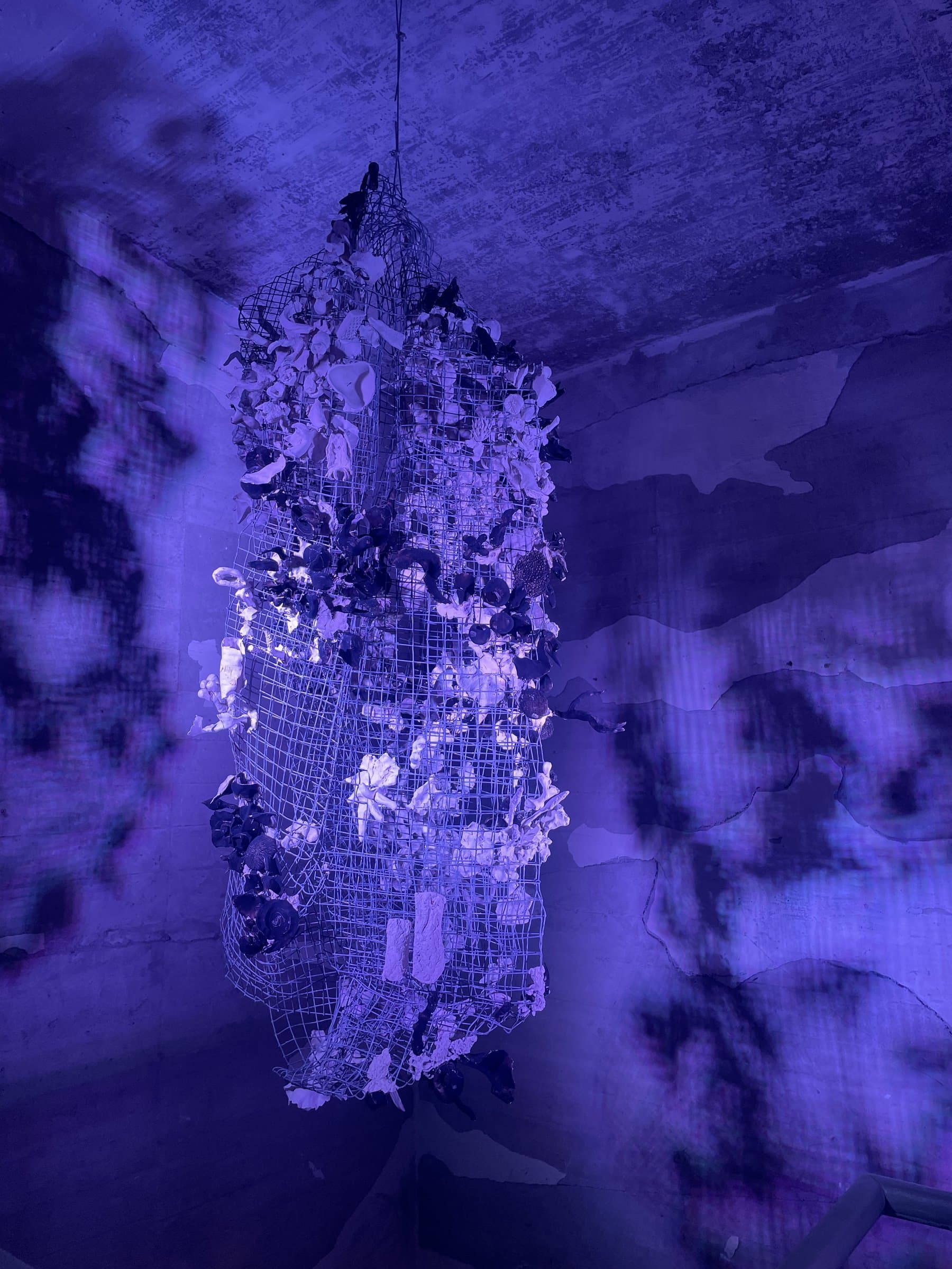 The finished sculpture hanging from the ceiling of the power plant, a wire mesh form covered in white and dark ceramic pieces, lit blue-violet