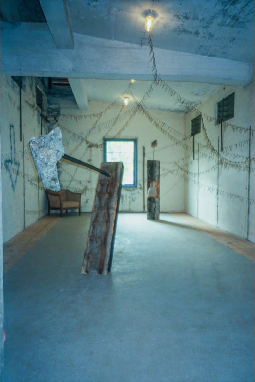 Wide view of the room: a leaning wooden plank and stone sculpture, a wired chandelier-like web of nails strung across the ceiling and walls, a chair by the window