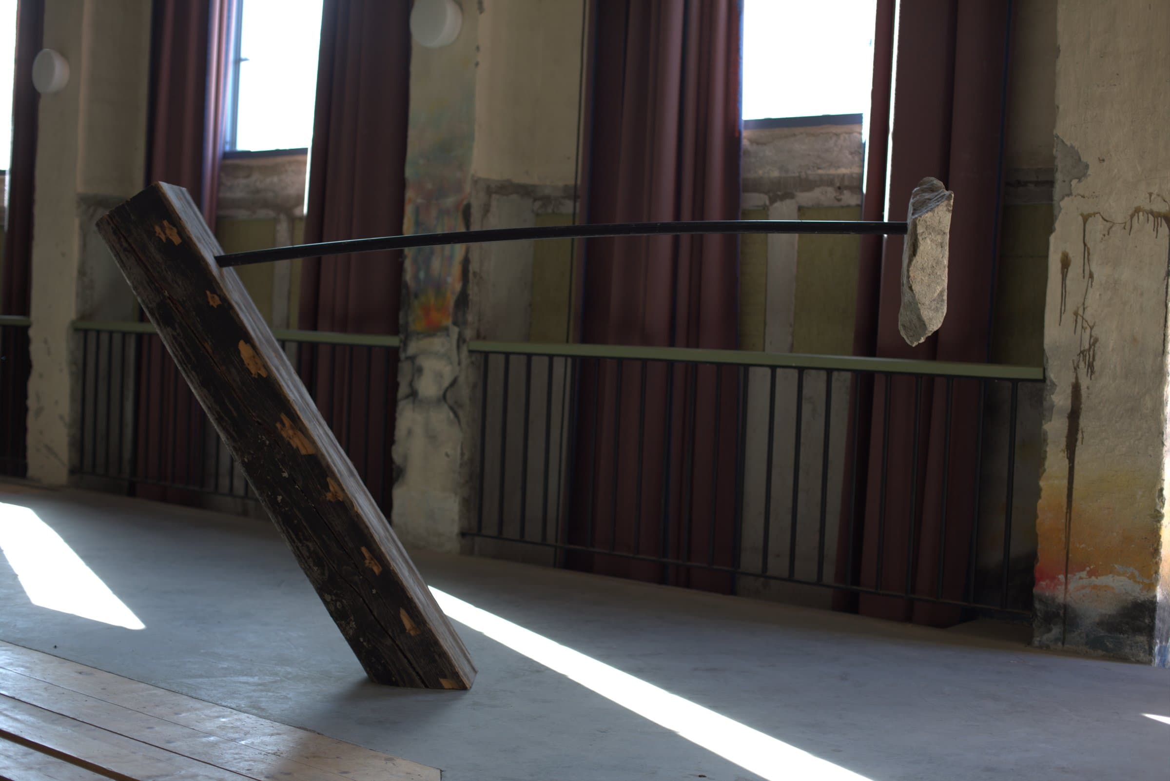 A wooden plank leaning at an angle, propped up by a curved black metal rod balancing a stone at its far end, in a sunlit room with red curtains