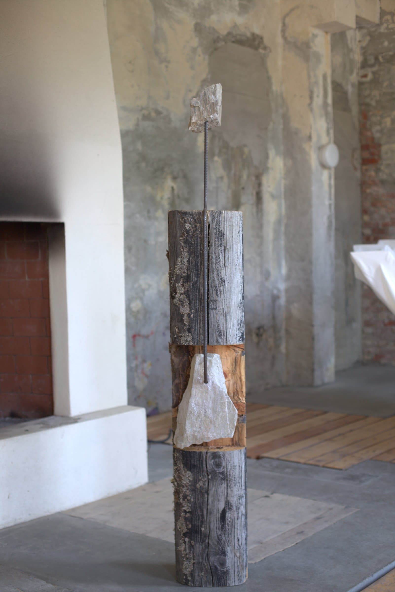 A tall wooden log column with a metal rod through its center, holding one stone wedged partway up and another balanced on top
