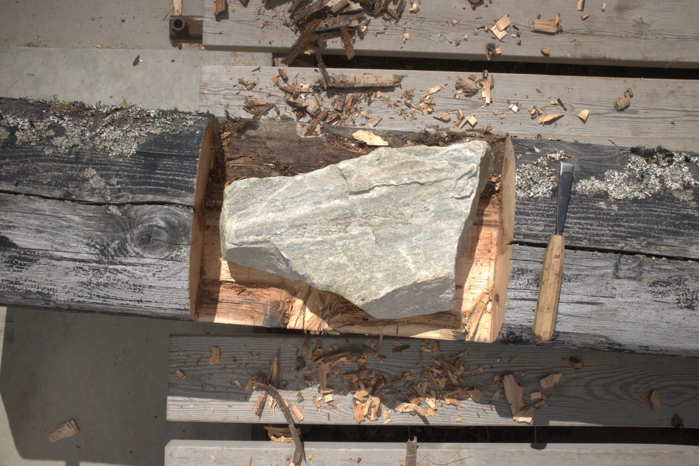 Overhead view of a stone being fitted into a carved notch in a wooden beam, wood chips scattered around it and a chisel resting alongside