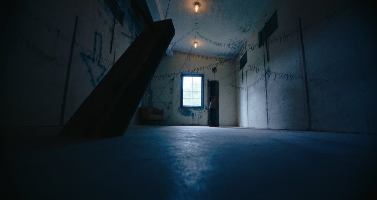 Wide interior view of the leaning plank and stone sculpture, a wired web of nails across the ceiling and walls, a chair by the window