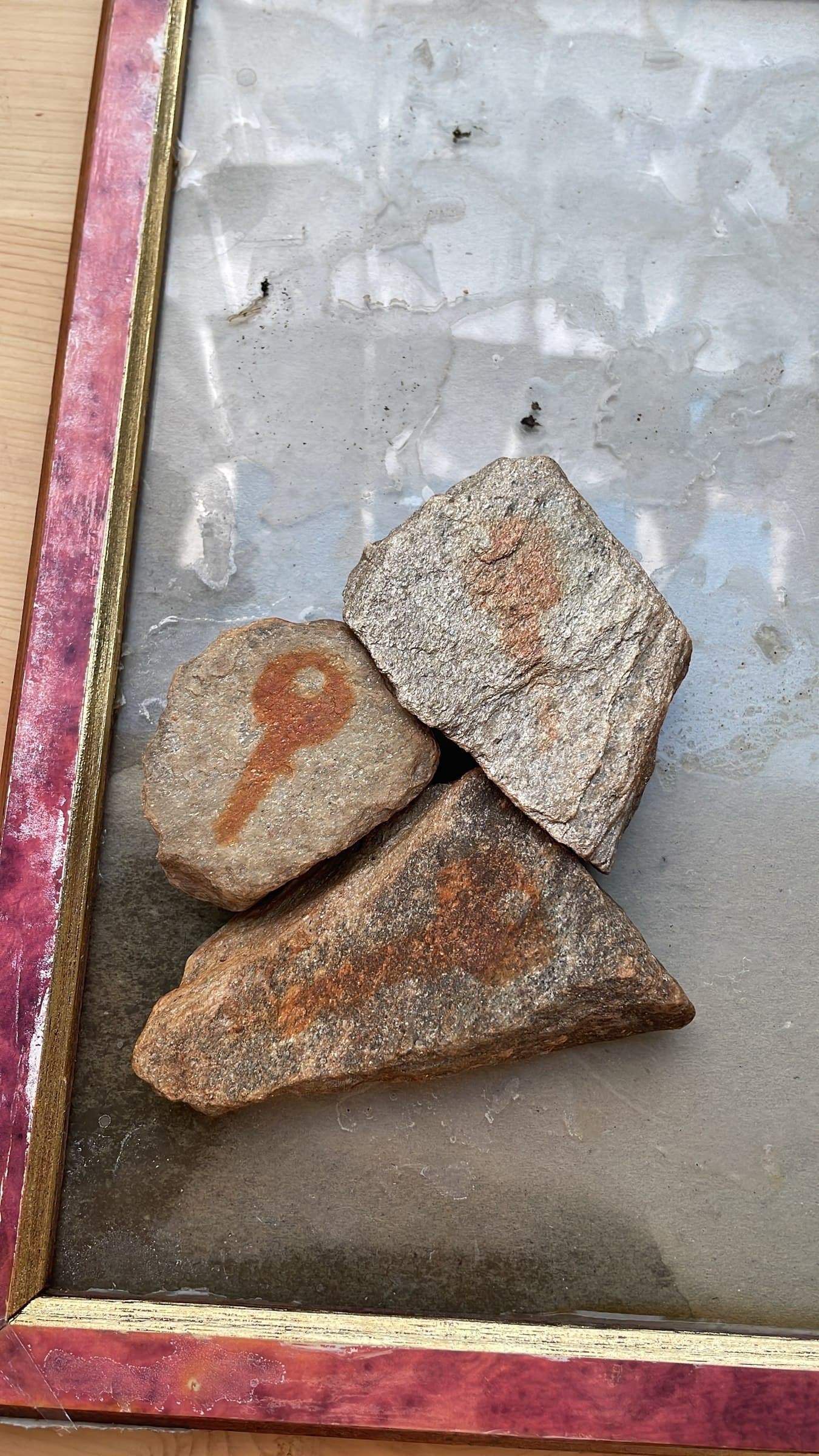 Three stones arranged in the corner of a red and gold picture frame, one marked with a small key shape and the other two with looser rust-red smudges