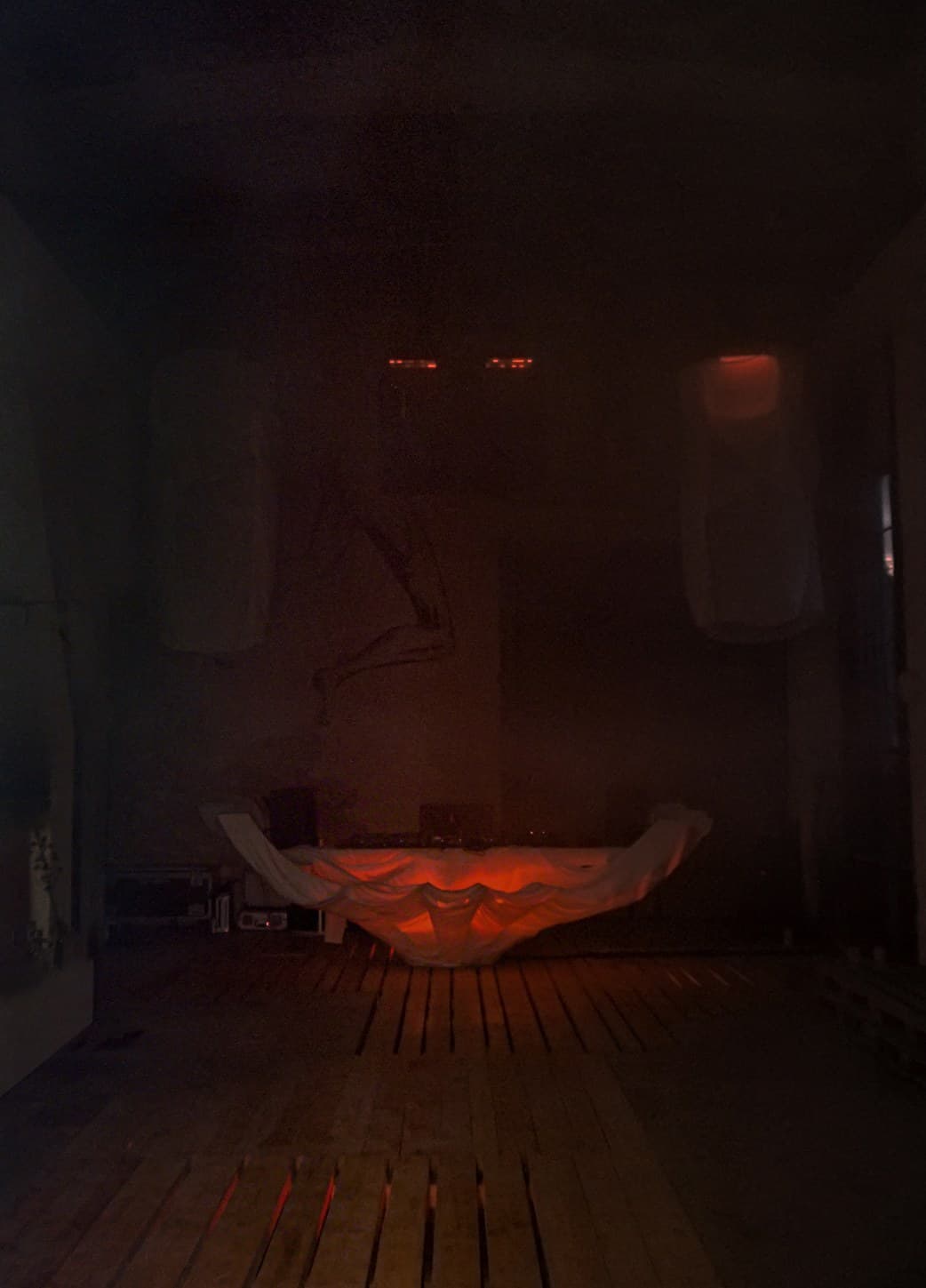 The draped structure lit from within in deep red, alone in the dark machine hall, smoke hanging in the air above the wooden floor