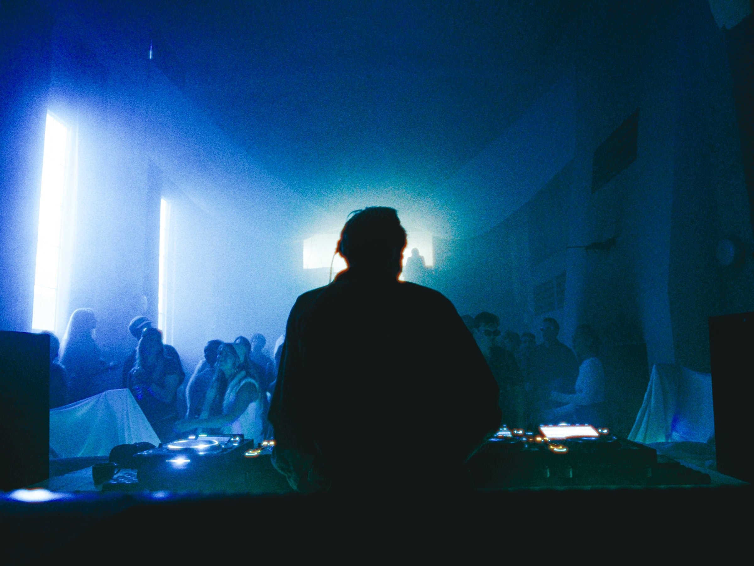 A DJ silhouetted at the decks in blue light, a crowd gathered by glowing windows behind them