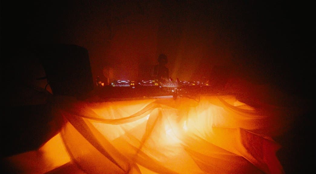 The same DJ booth lit warm orange, draped fabric glowing beside the decks