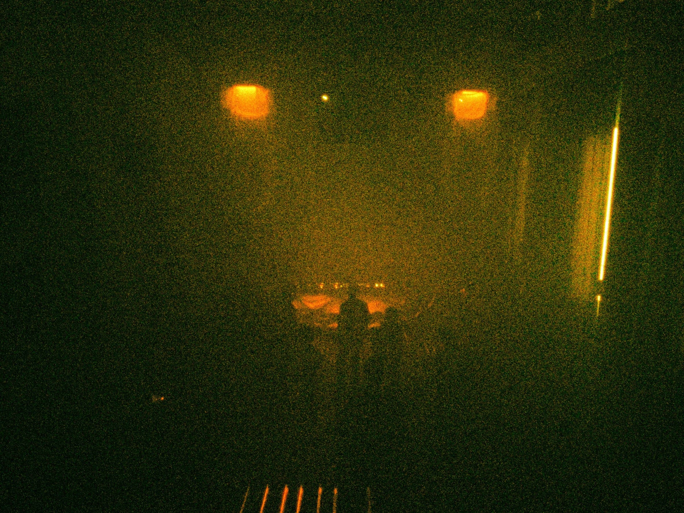 A grainy night shot with two glowing lights, a vertical light streak, and a draped structure visible in the distance