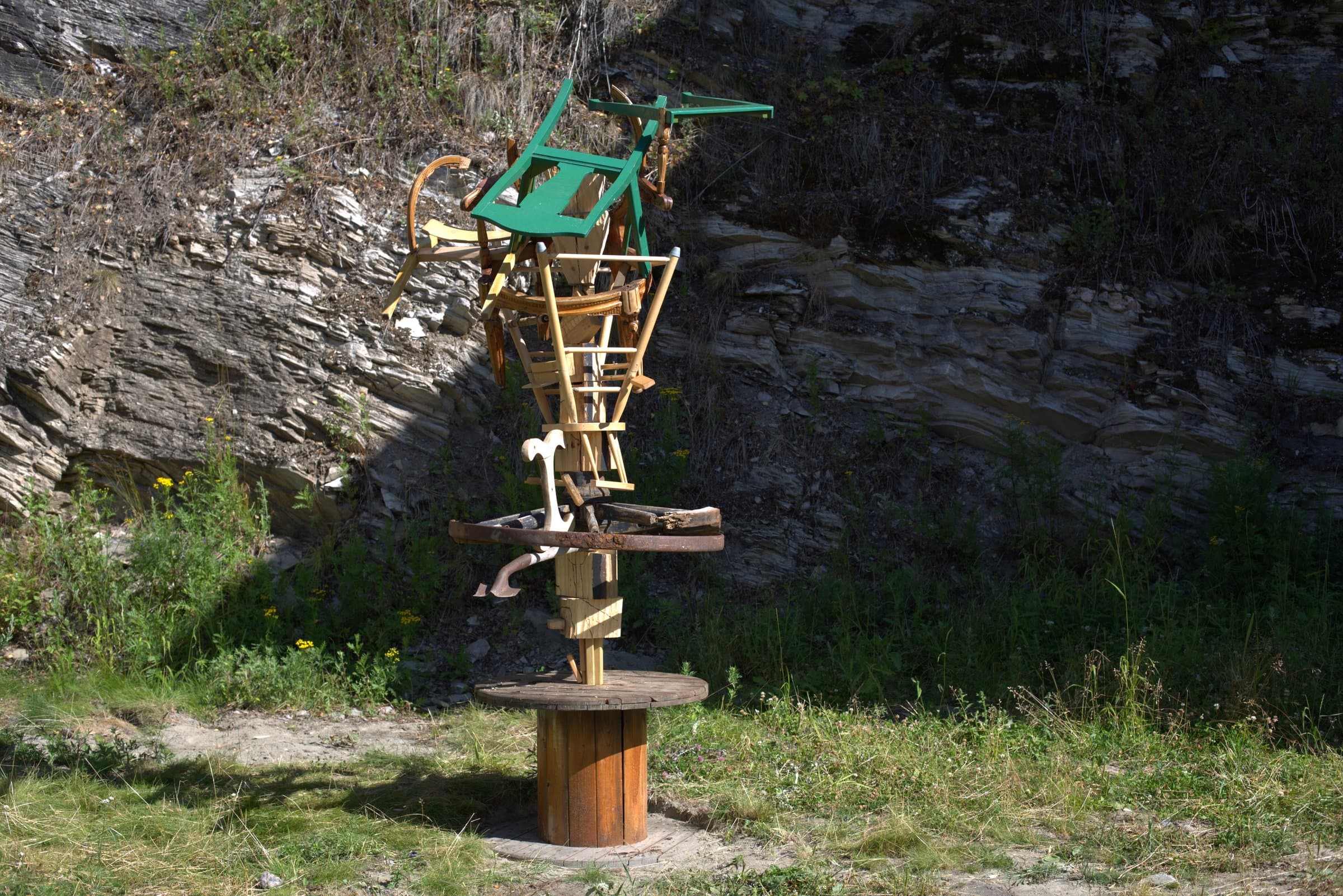 A totem-like stack of chairs and wood, a green painted chair on top, standing on a wooden cable spool against a rocky cliff face