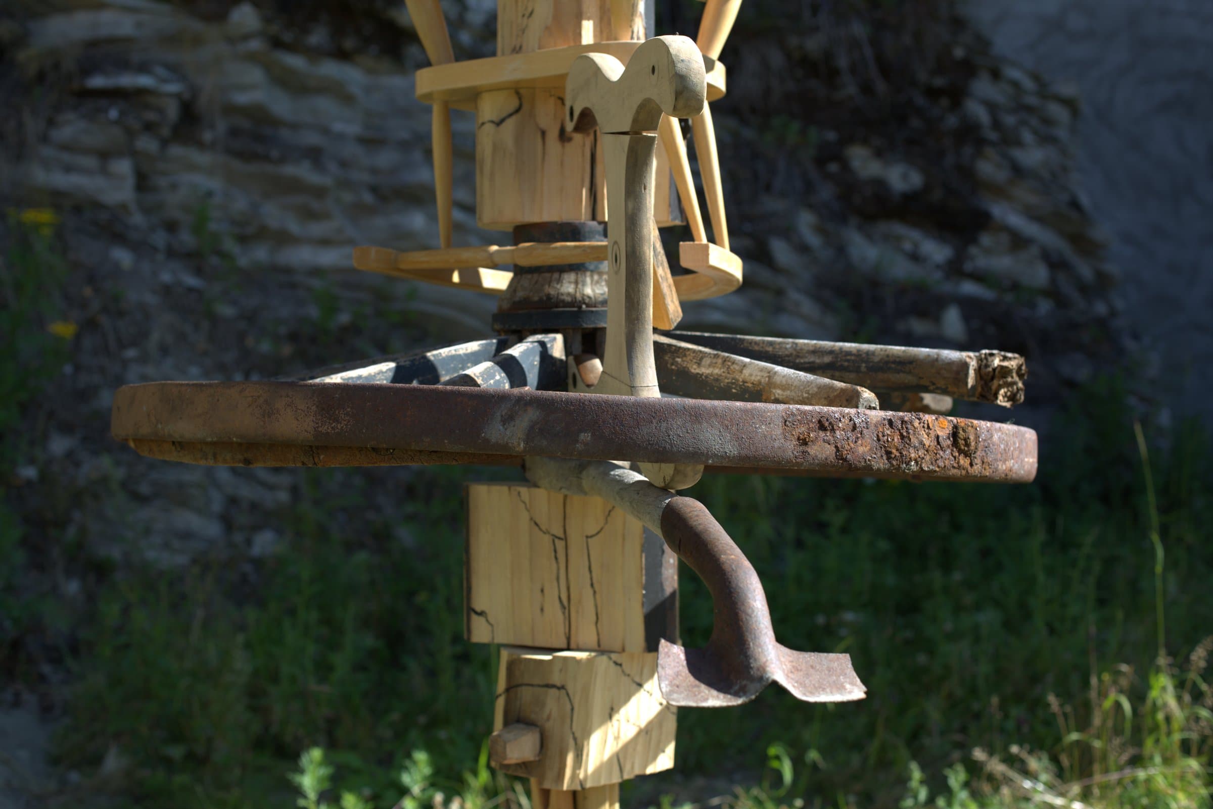 Close-up of the middle of the sculpture, a carved wooden horse-head shape, a rusted metal wheel, and an old hooked tool