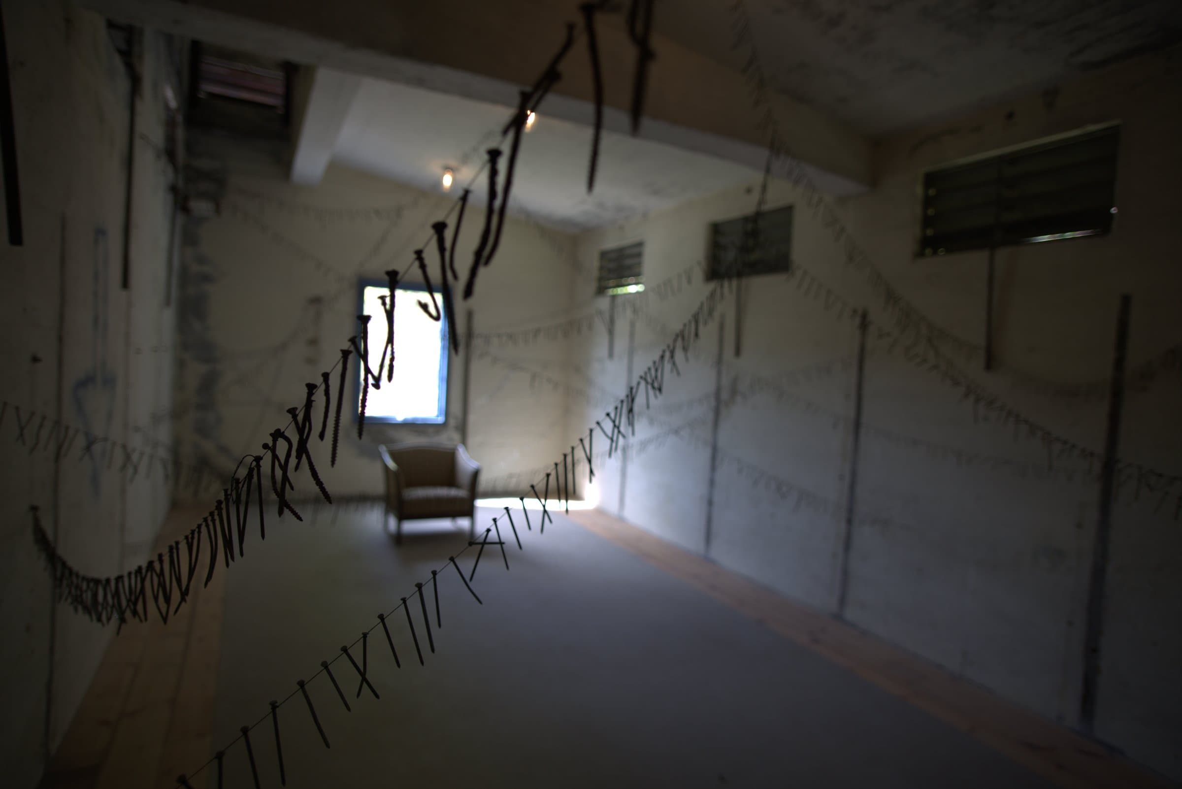 A concrete room strung with wires of hanging nails and screws in a zigzag pattern, an armchair and a sunlit window in the background