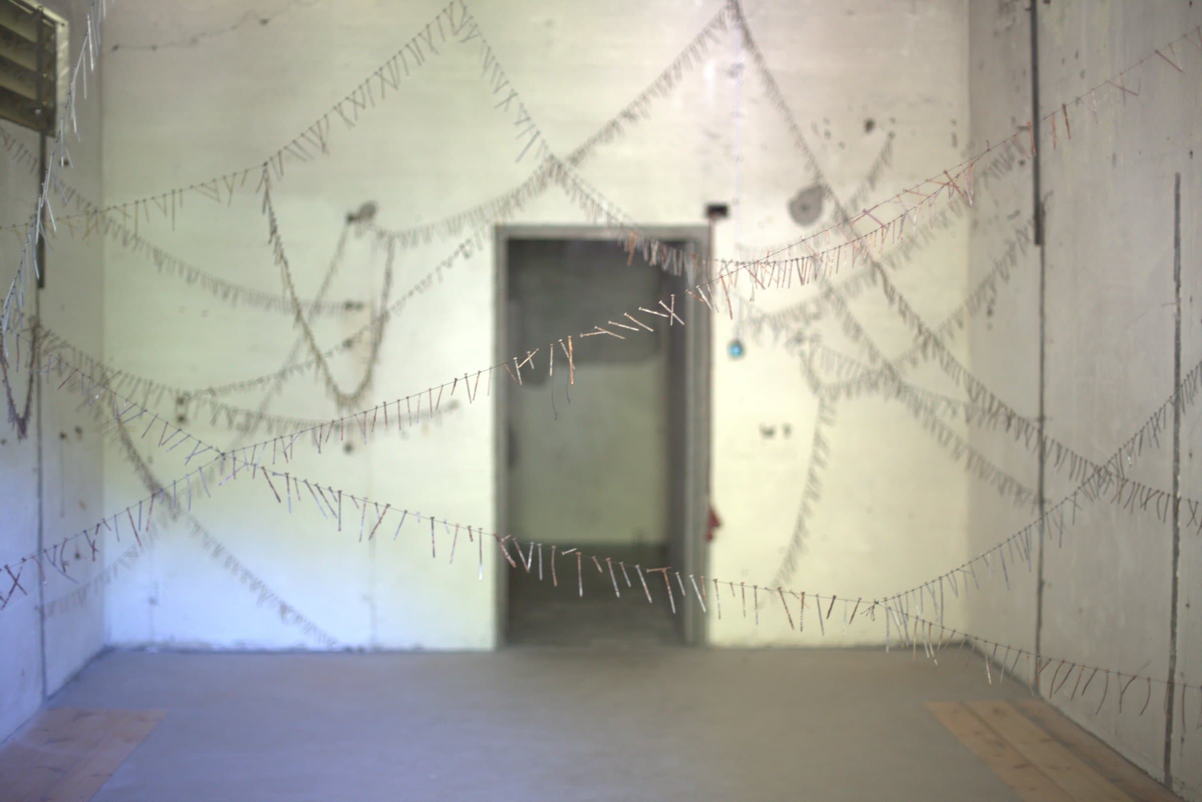 Wide view of the installation filling the room, diagonal wires of nails crisscrossing the walls around a central doorway