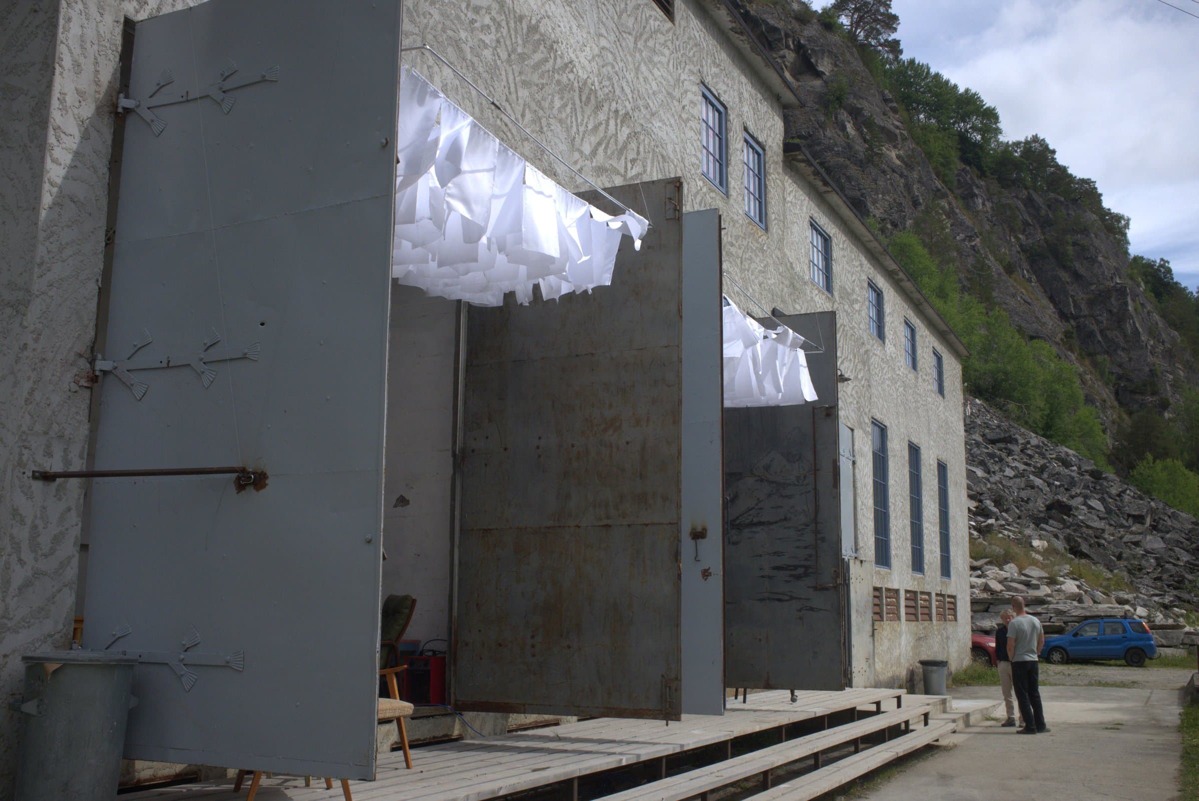 The finished installation: pale fabric flags hanging in the open doorway under the roof edge of the power plant building, two people looking on from outside