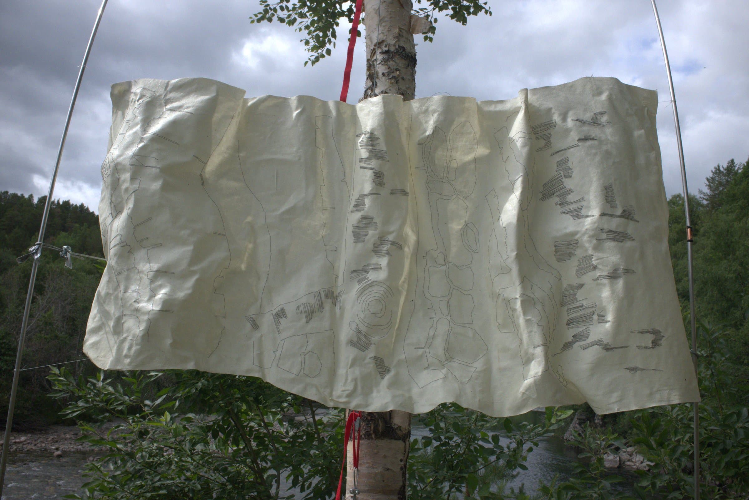 The drawn fabric panel strung between metal poles by the riverbank, seen from below against a cloudy sky