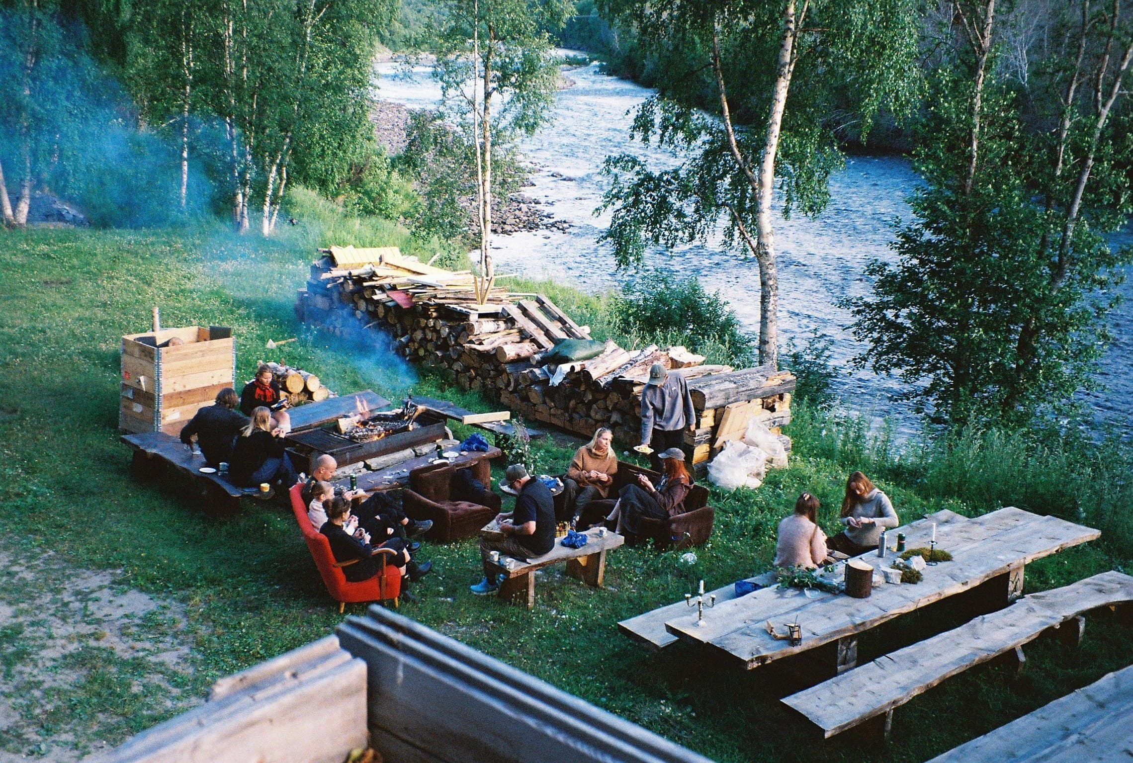 An overhead view of a group gathered around a fire pit and long wooden tables on the riverbank at dusk, eating and talking, with a stack of firewood and birch trees around them.