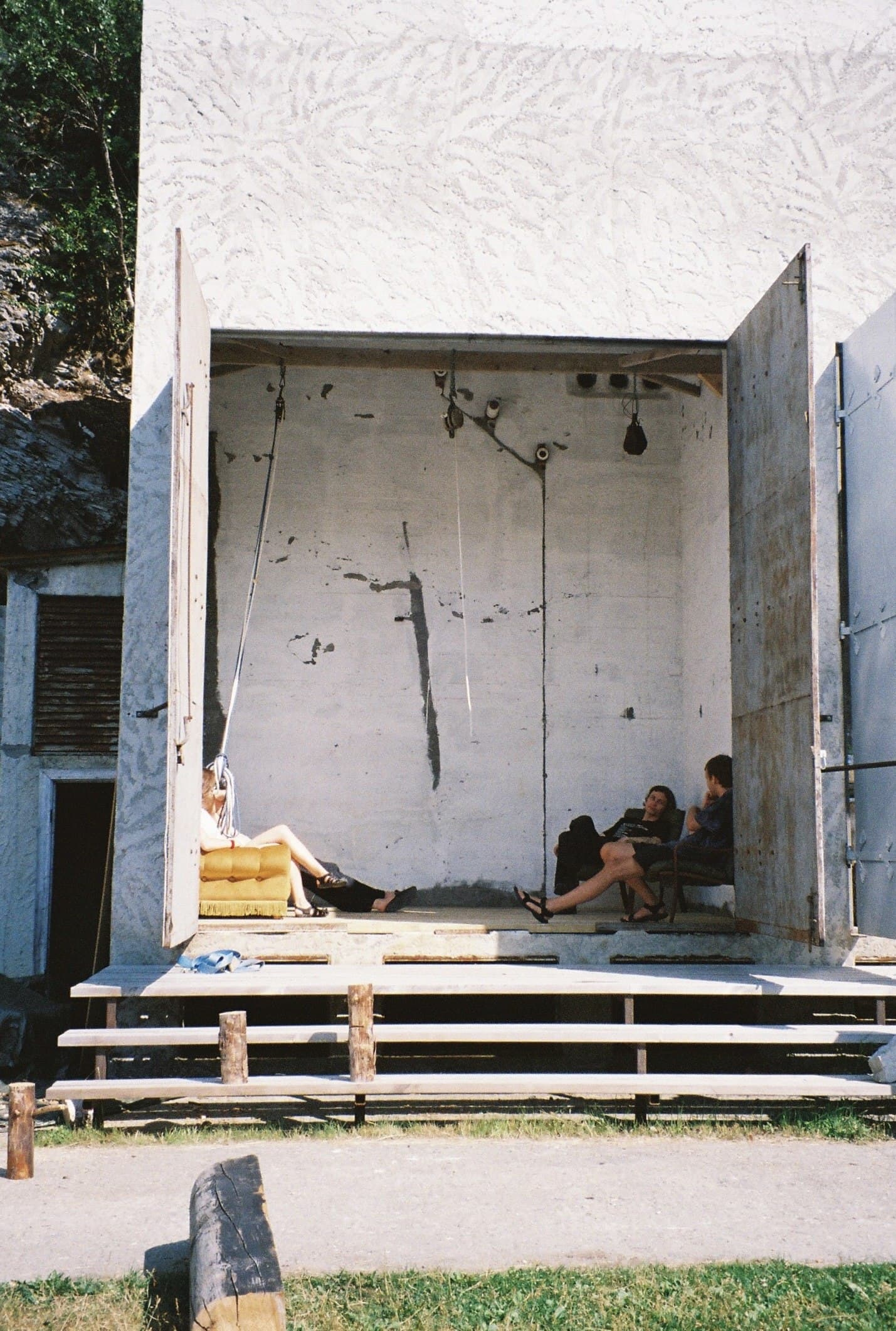 The building's raw concrete machine hall, its large double doors open, with people sitting and talking on a sofa and chairs inside.
