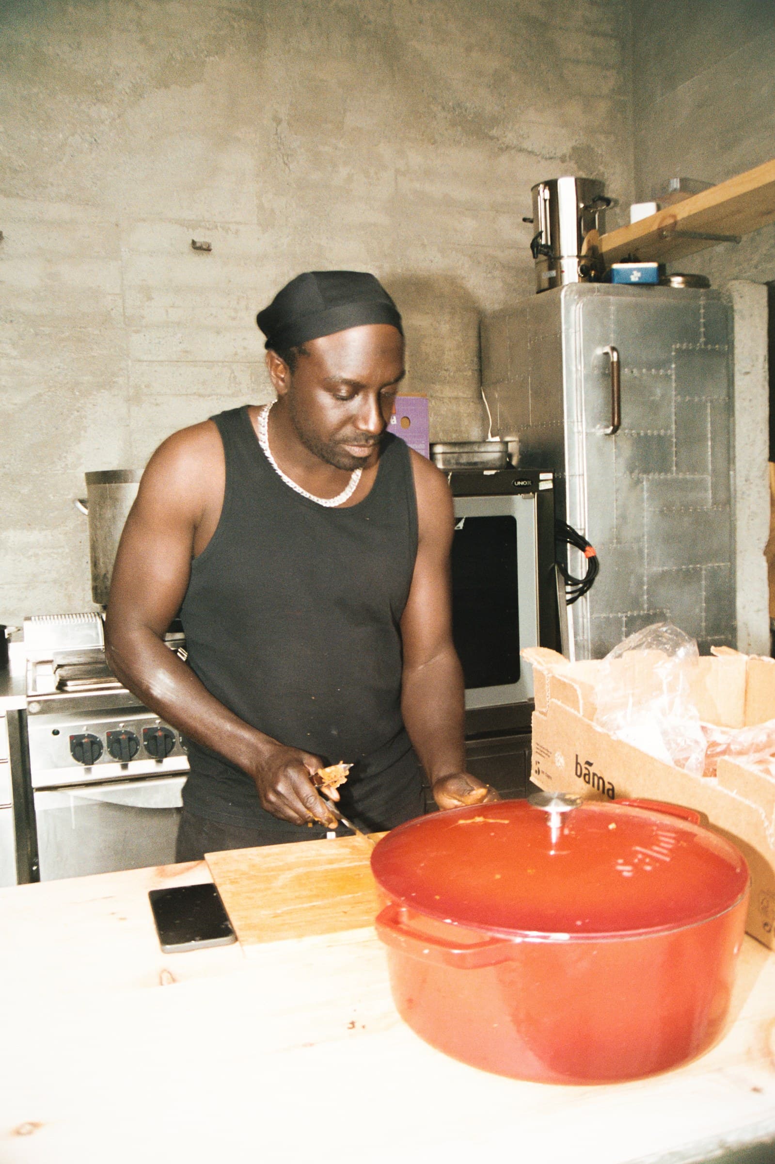 Someone cooking in the kitchen at a plywood counter with a large red pot in front of them, concrete walls and a steel fridge behind.