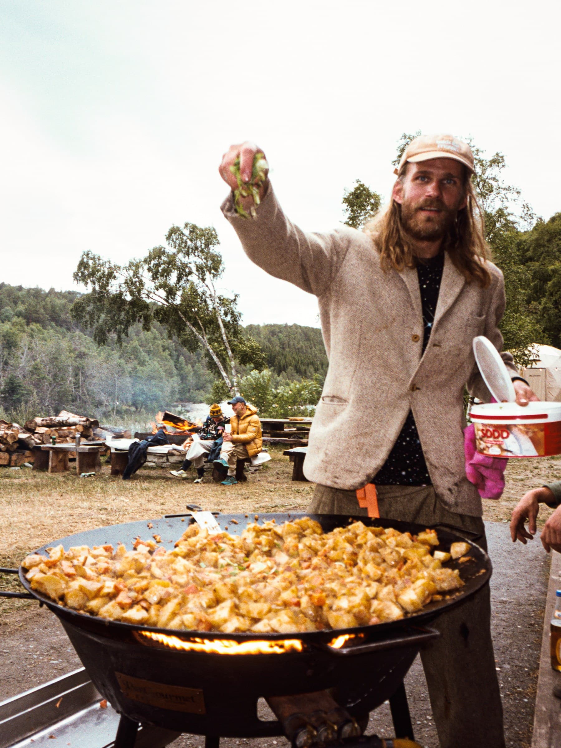 Someone scattering herbs over a huge pan of potatoes cooking over a flame outdoors, with the fire pit, stacked firewood, birch trees and the river behind them.