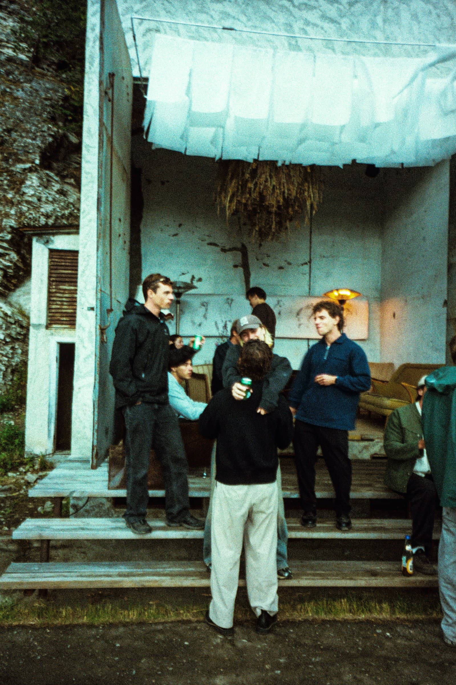 People standing and sitting with drinks on the wooden steps of an outbuilding under a stretched fabric canopy, the rock face rising behind it.