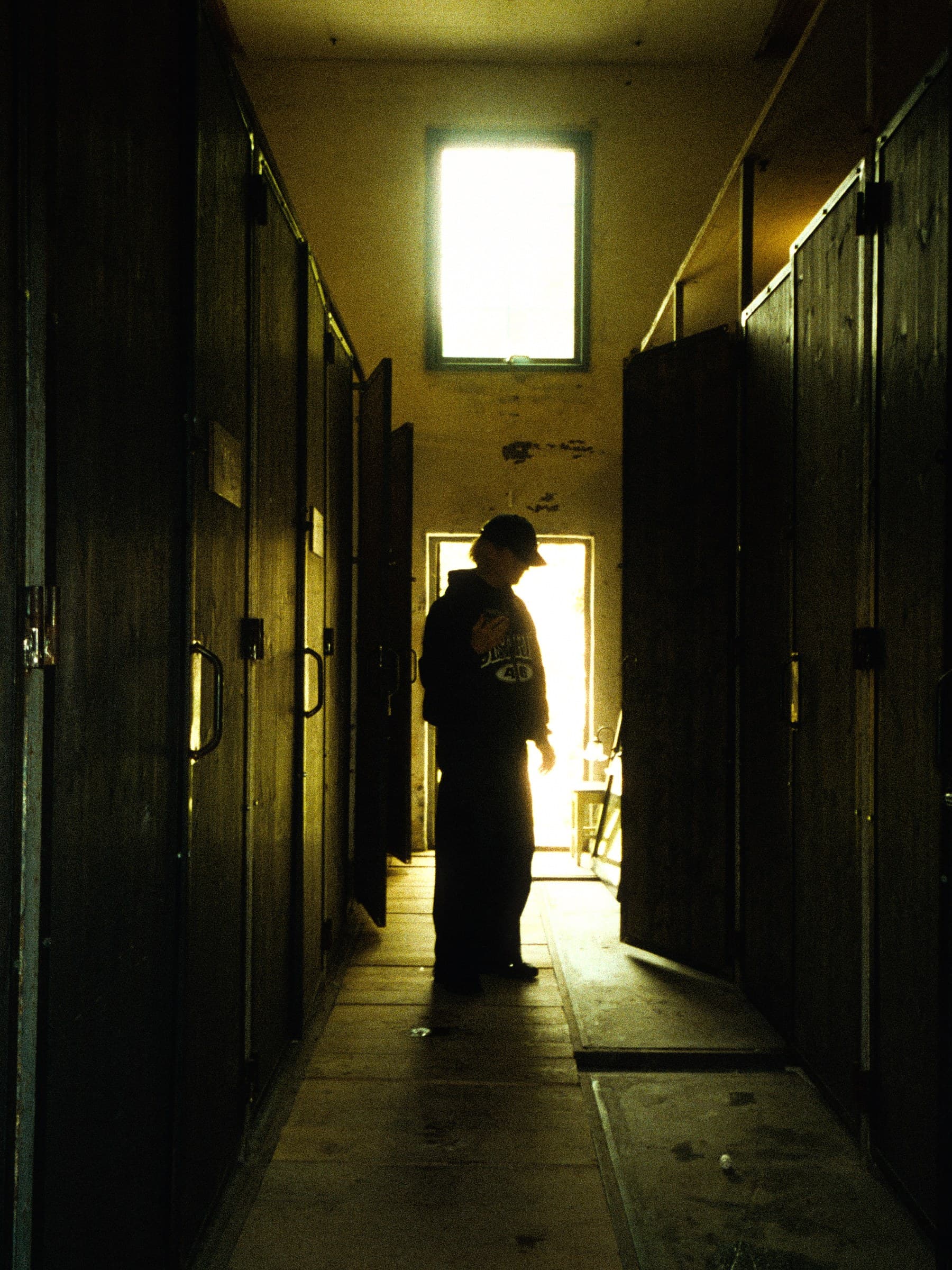 A dim corridor lined with wooden bedroom doors, someone standing silhouetted against the bright doorway at the far end.