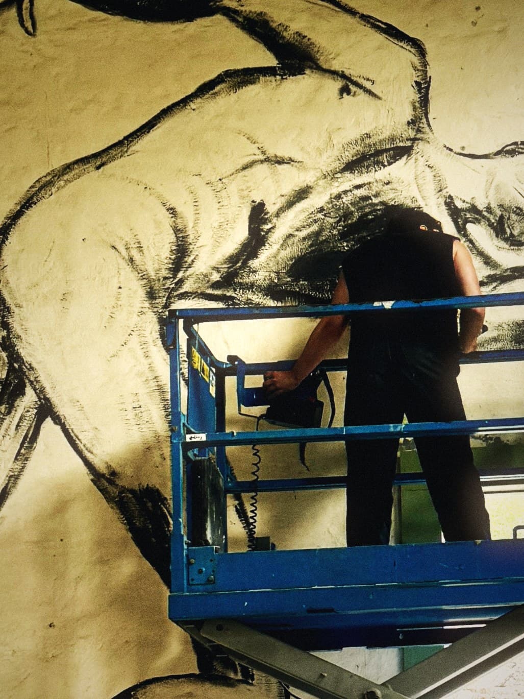 An artist on a raised scissor lift, working on an enormous charcoal drawing of an animal that covers the whole wall behind them.