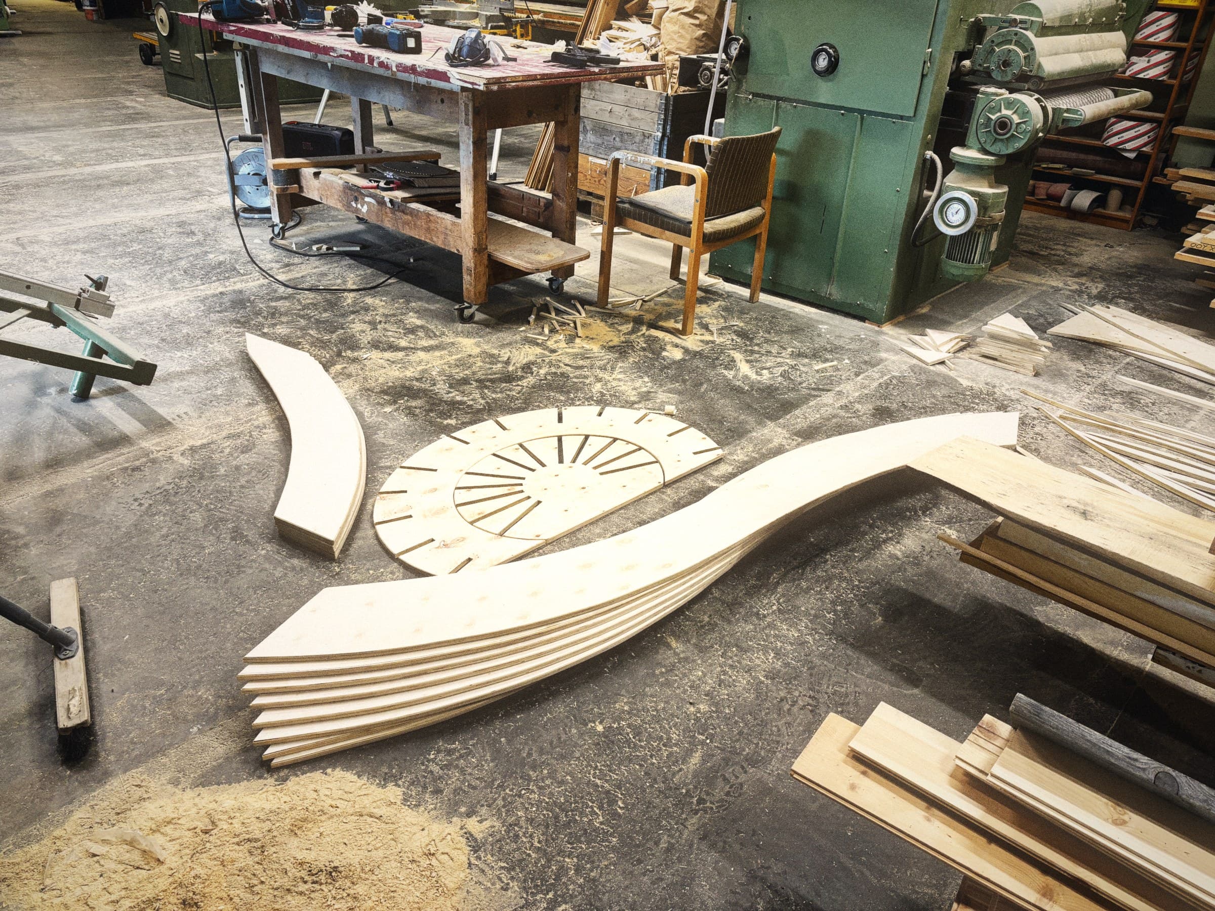 The wood workshop: curved laminated plywood forms and a slotted disc laid out on the sawdust-covered floor, with a workbench, a bandsaw and a green industrial planer behind them.