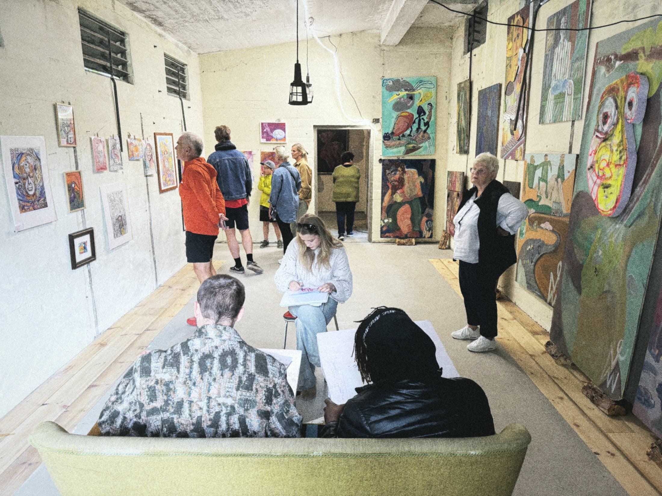 Visitors at an exhibition on the production floor, paintings covering the walls, two people seated on a sofa.
