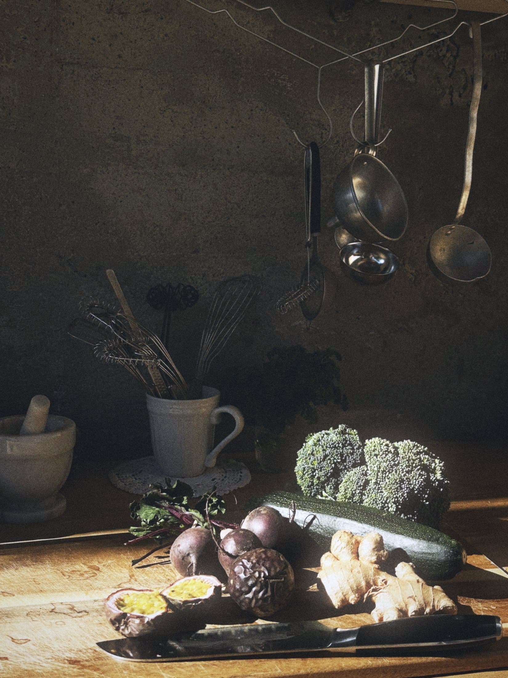 Vegetables and ginger on a wooden counter, pans hanging from the wall above.