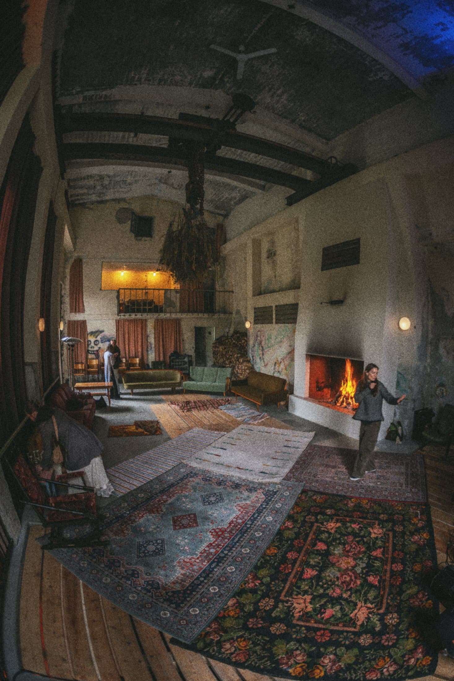 The machine hall seen wide, rugs on the wooden floor, a fire burning in the open fireplace and a person standing by it.