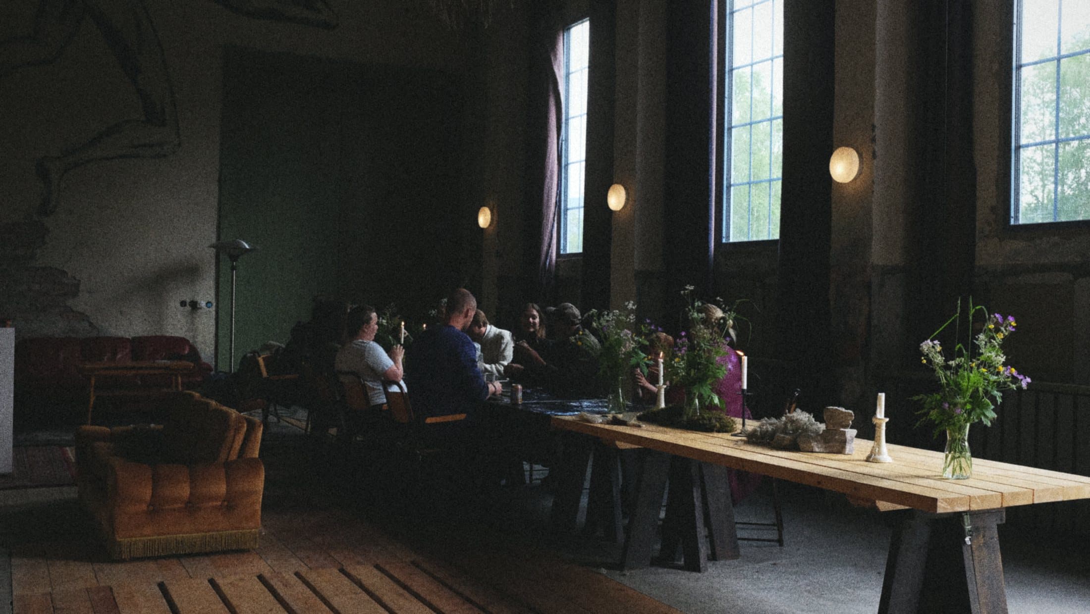 A long table set for dinner in the dim machine hall, tall windows and wall lamps along one side.