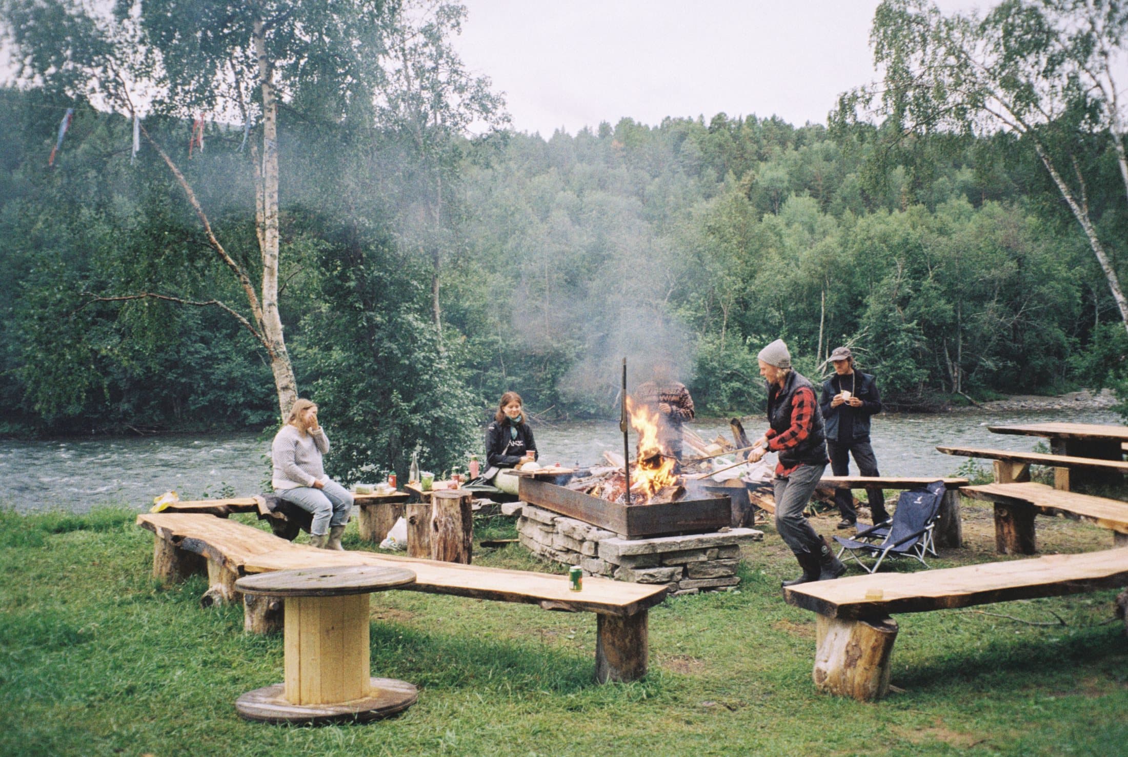 People around the fire pit by the river, sitting on log benches arranged in a ring, smoke rising into the birches.