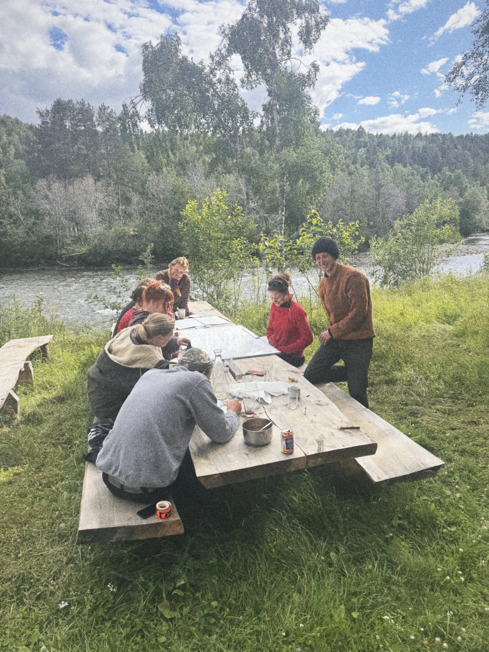 A group working at a long wooden table on the grass by the river.